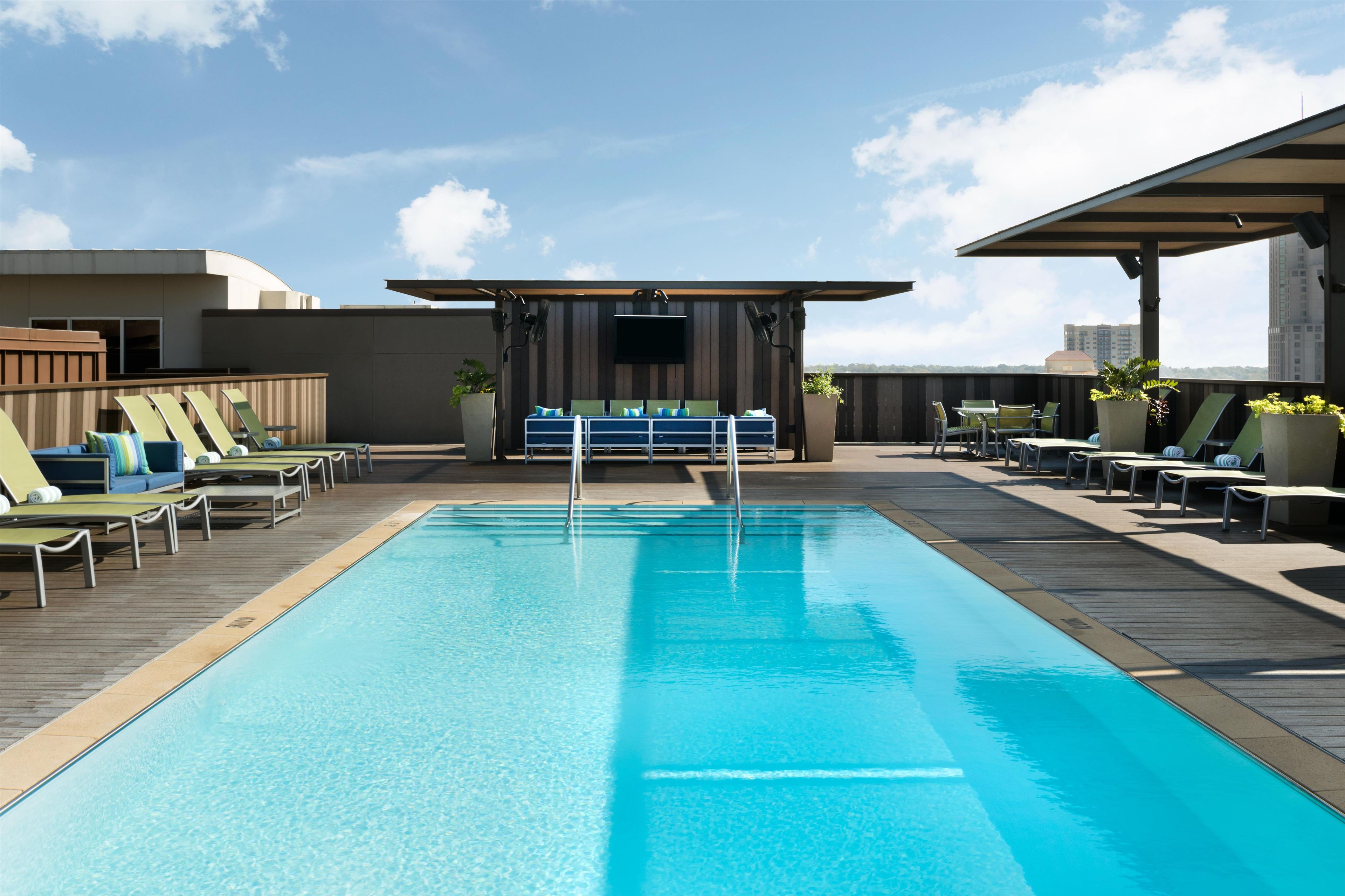 Una piscina en la azotea con sillones de descanso, una zona de bar y vistas del horizonte de la ciudad.