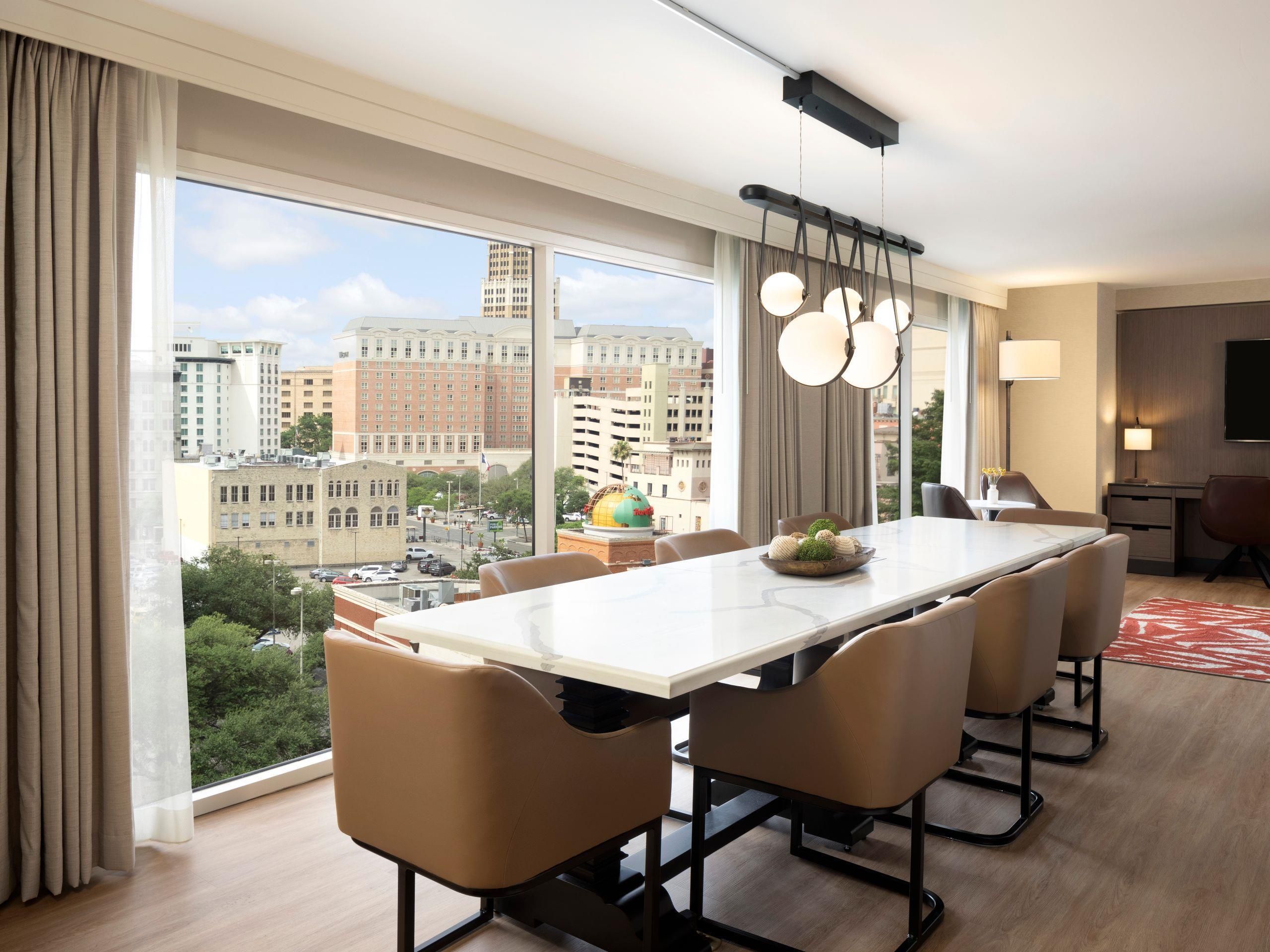 A modern hotel room with a large dining table, comfortable chairs, a city view, and a flat-screen TV.