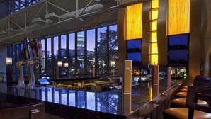 Un área de bar moderna con techo alto y ventanas amplias, una barra con taburetes y vistas a un paisaje urbano al atardecer.