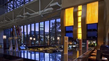 A modern bar area with a high ceiling and large windows, a counter with stools, and a view of a cityscape at dusk.