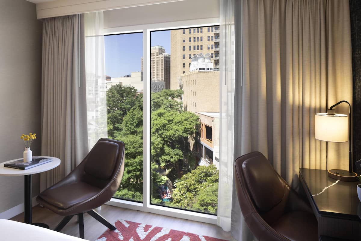 Habitación de hotel con un ventanal grande, dos sillones de cuero, una mesita con un florero y un libro, y una lámpara en una mesita de noche.