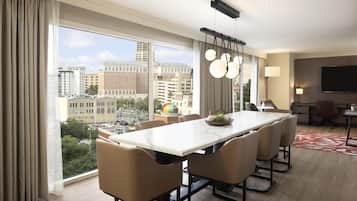 A modern hotel room with a large dining table, comfortable seating, a flat-screen TV, and a city view.