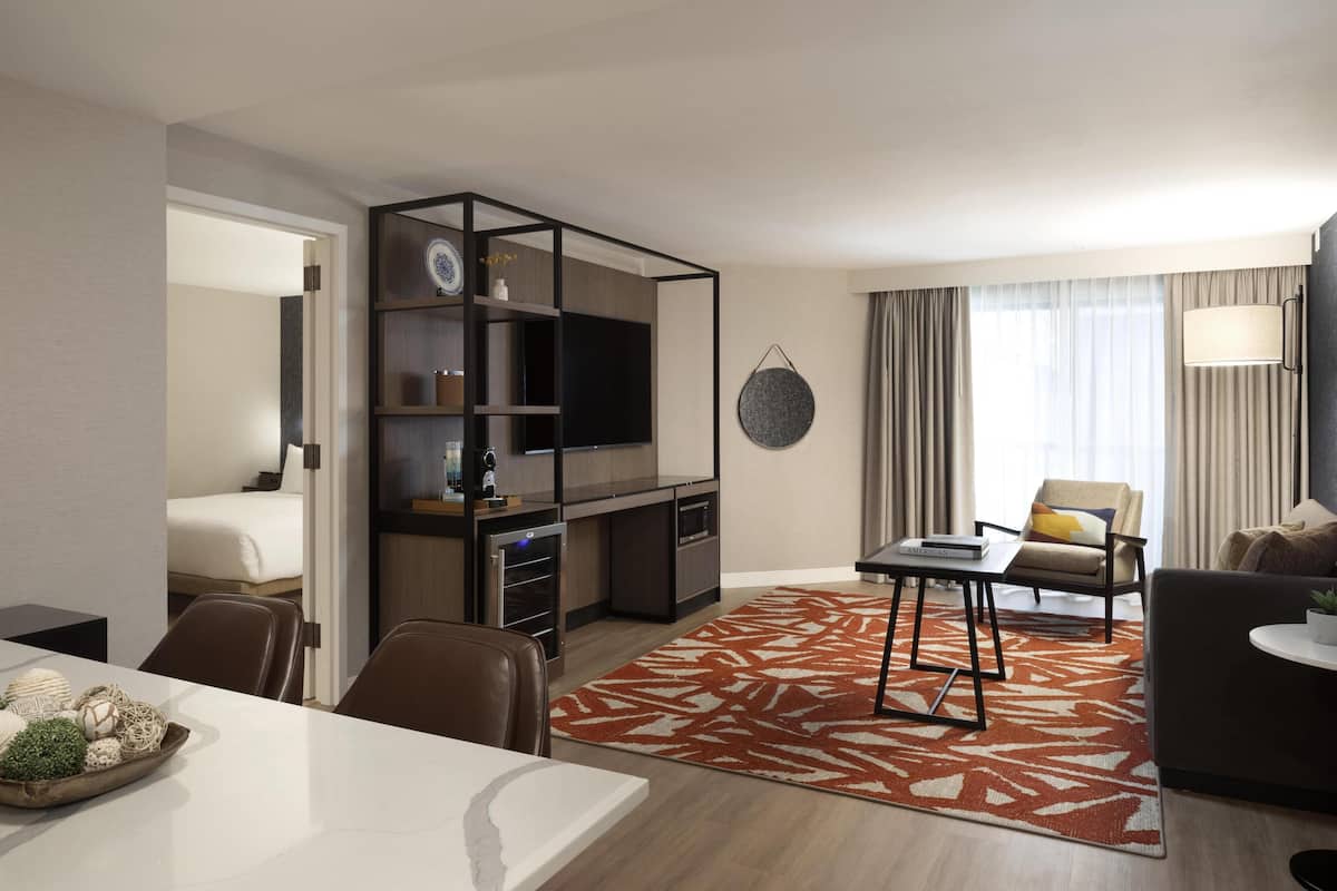 Habitación de hotel con zona de comedor, sofá, televisión, estantería y ventana con cortinas.