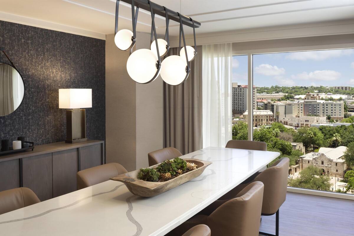 Un comedor moderno con una mesa grande, sillas, una lámpara colgante, un espejo y vistas de la ciudad.
