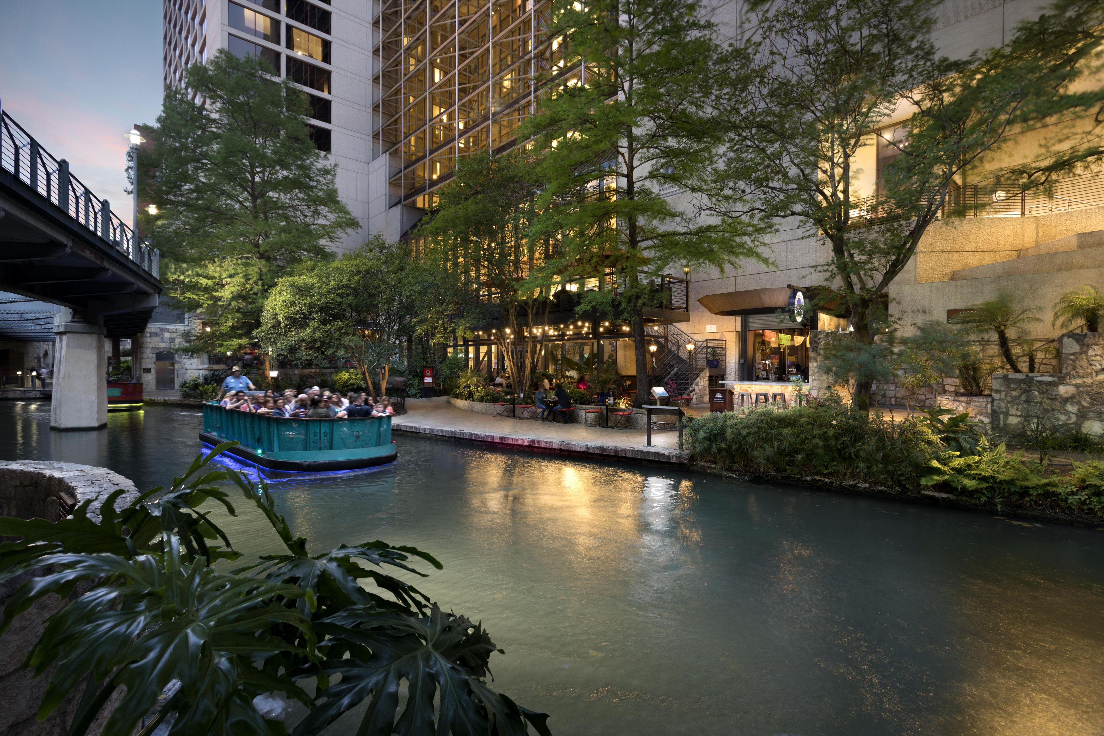 A riverside walkway with a boat ride, surrounded by greenery and modern buildings.