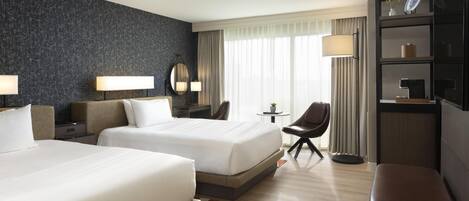 A hotel room with two beds, a desk, a chair, a small table, a TV, and a bookshelf.