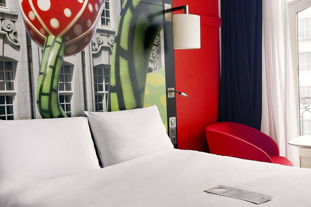 A hotel room with a bed, a red chair, a wall with a large abstract painting, and a window with curtains.