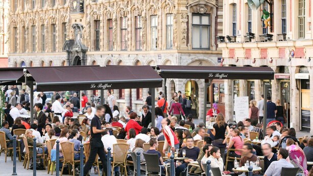 Un café al aire libre con mesas en la terraza, un camarero y mucha gente.