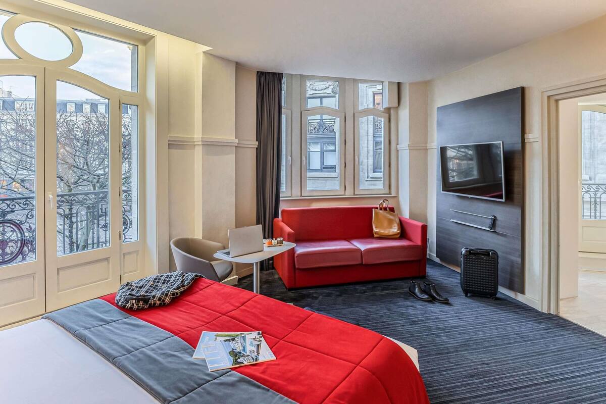 A hotel room with a bed, a red sofa, a TV, and a window with a view of buildings.