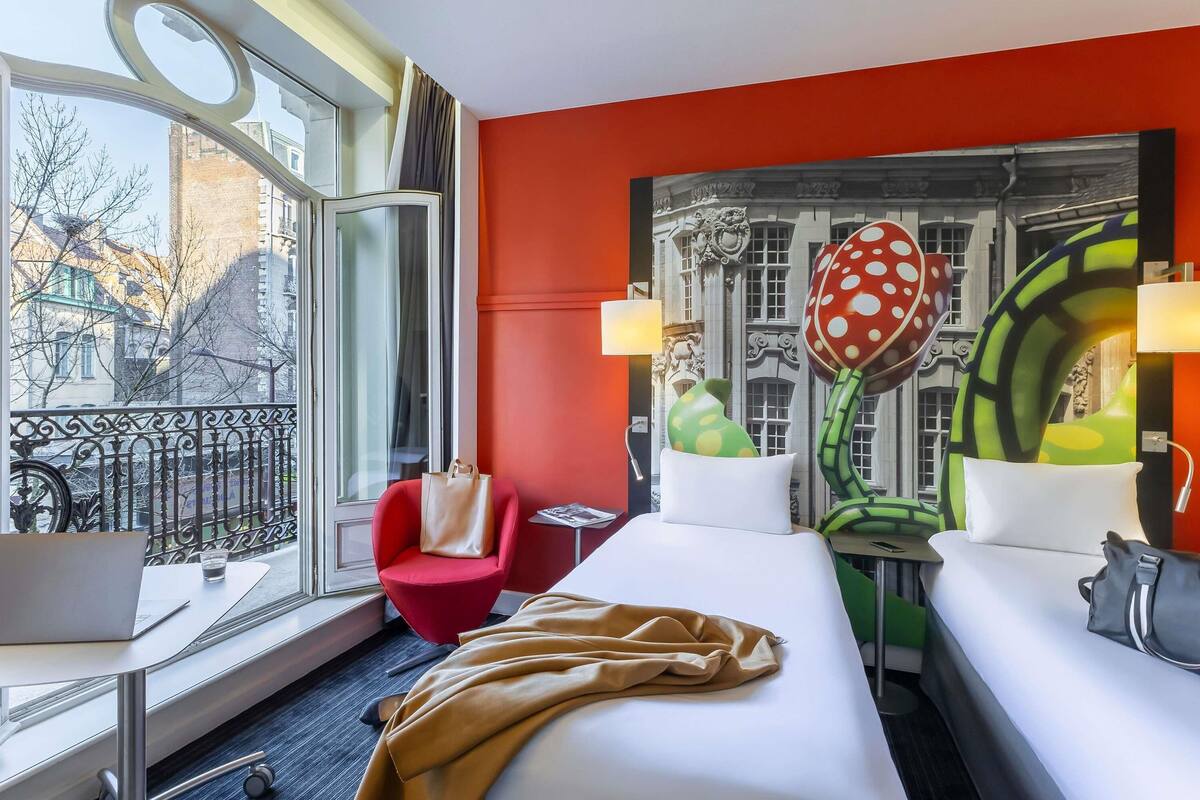 A hotel room with a large window, a red chair, a bed with a beige blanket, and a unique wall decoration featuring a mushroom and a building facade.