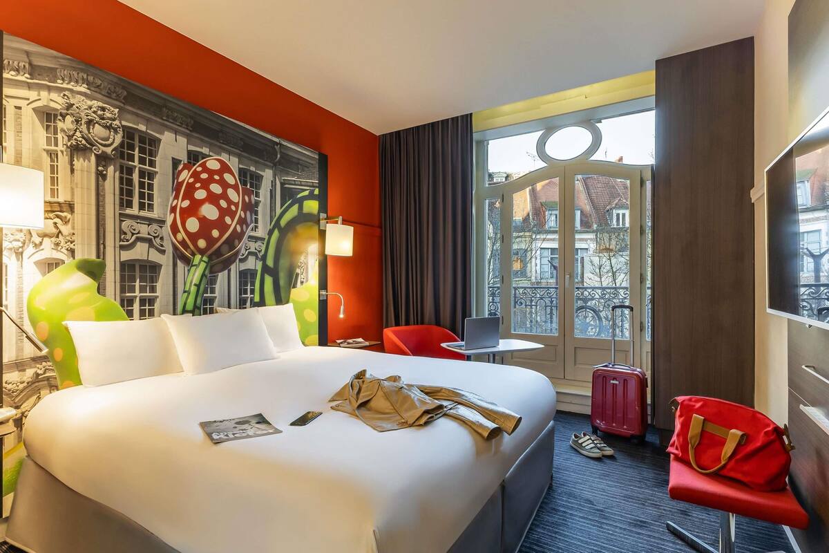 A hotel room with a large bed, a red chair, a desk with a laptop, and a window with a view of buildings.