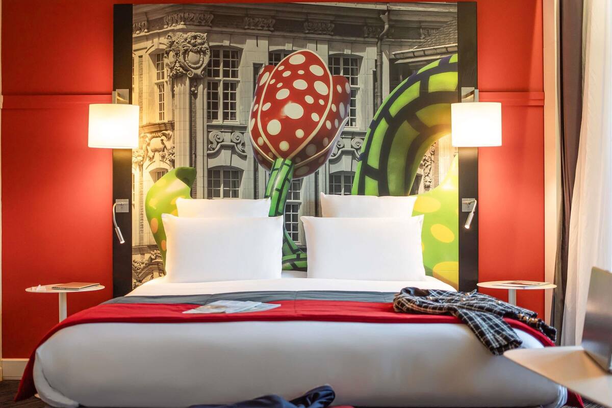 A hotel room with a large bed, two bedside tables, a red wall, and a large painting.