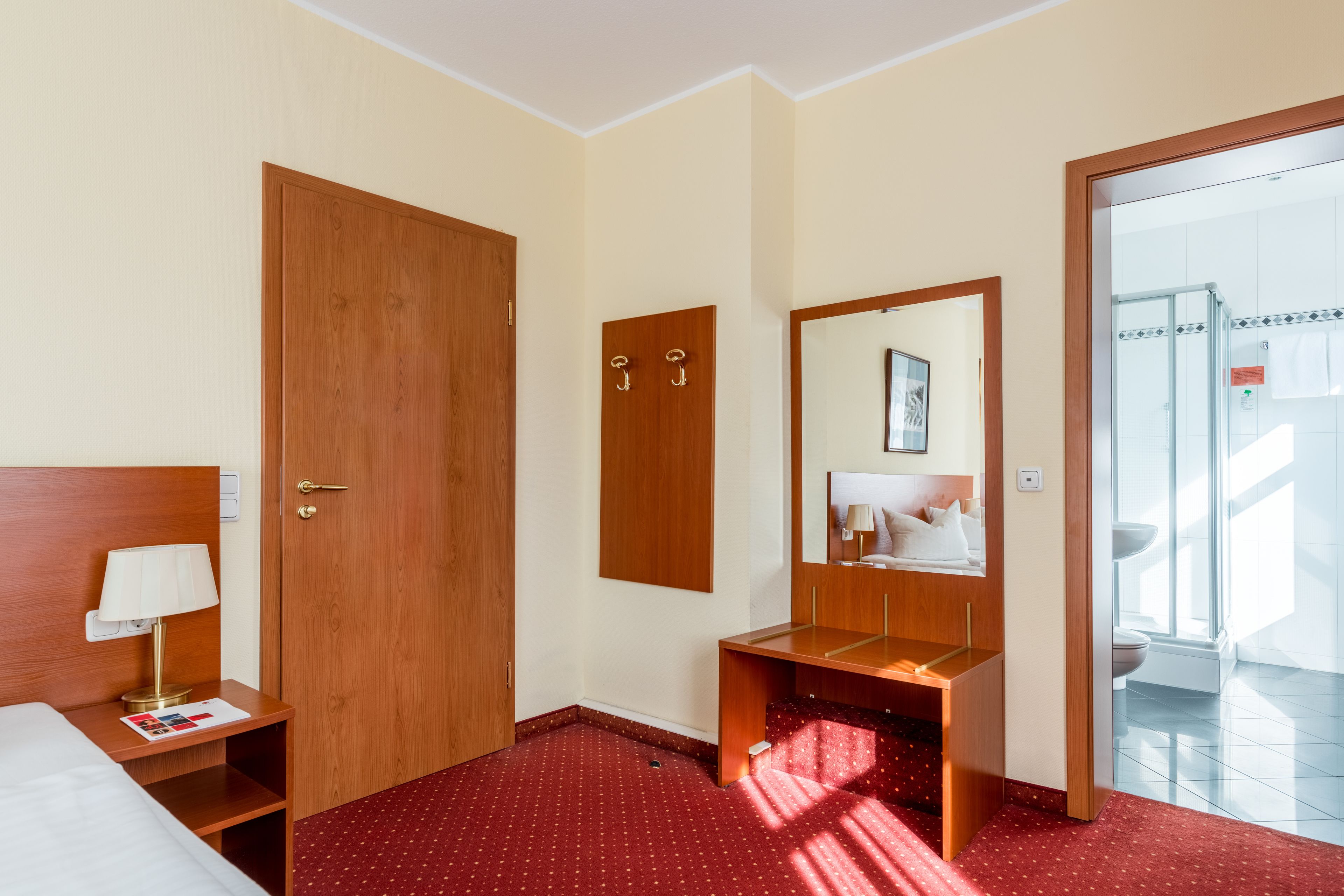 Quarto de hotel com porta de madeira, mesa de cabeceira com abajur, espelho e chão coberto por carpete vermelho.