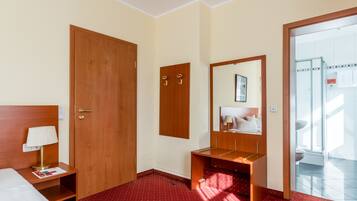 Quarto de hotel com porta de madeira, mesa de cabeceira com abajur, espelho e chão coberto por carpete vermelho.