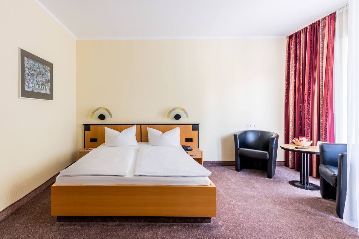 A hotel room with a large bed, a bedside table, a chair, a small table, and a window with curtains.