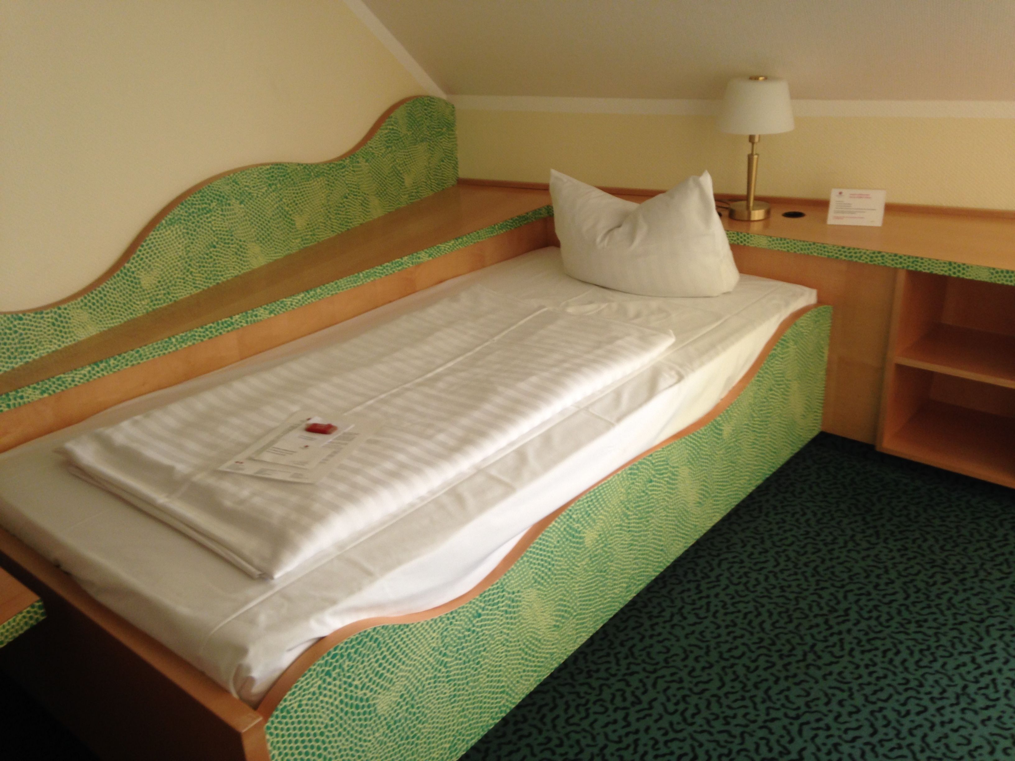 Una cama individual con tablero de cabecera y piecera estampados en verde, colchón blanco y una almohada.