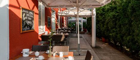 An outdoor cafe with wicker tables and chairs, umbrellas, and a red wall.