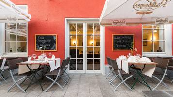 An outdoor cafe with tables set for dining, featuring a red exterior wall and white awnings.