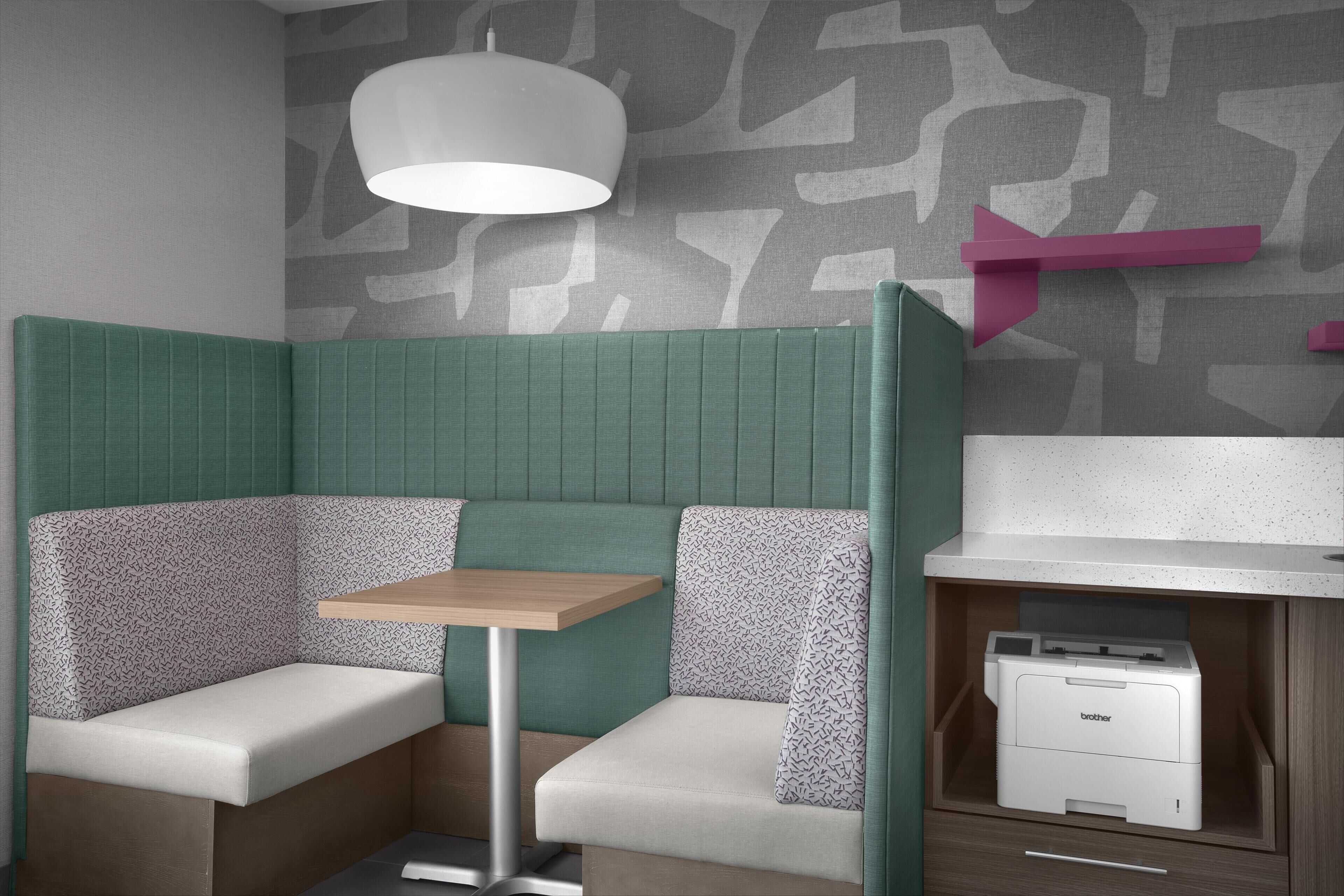 A corner booth with a table, patterned cushions, a ceiling lamp, a wall-mounted printer, and a patterned wall.