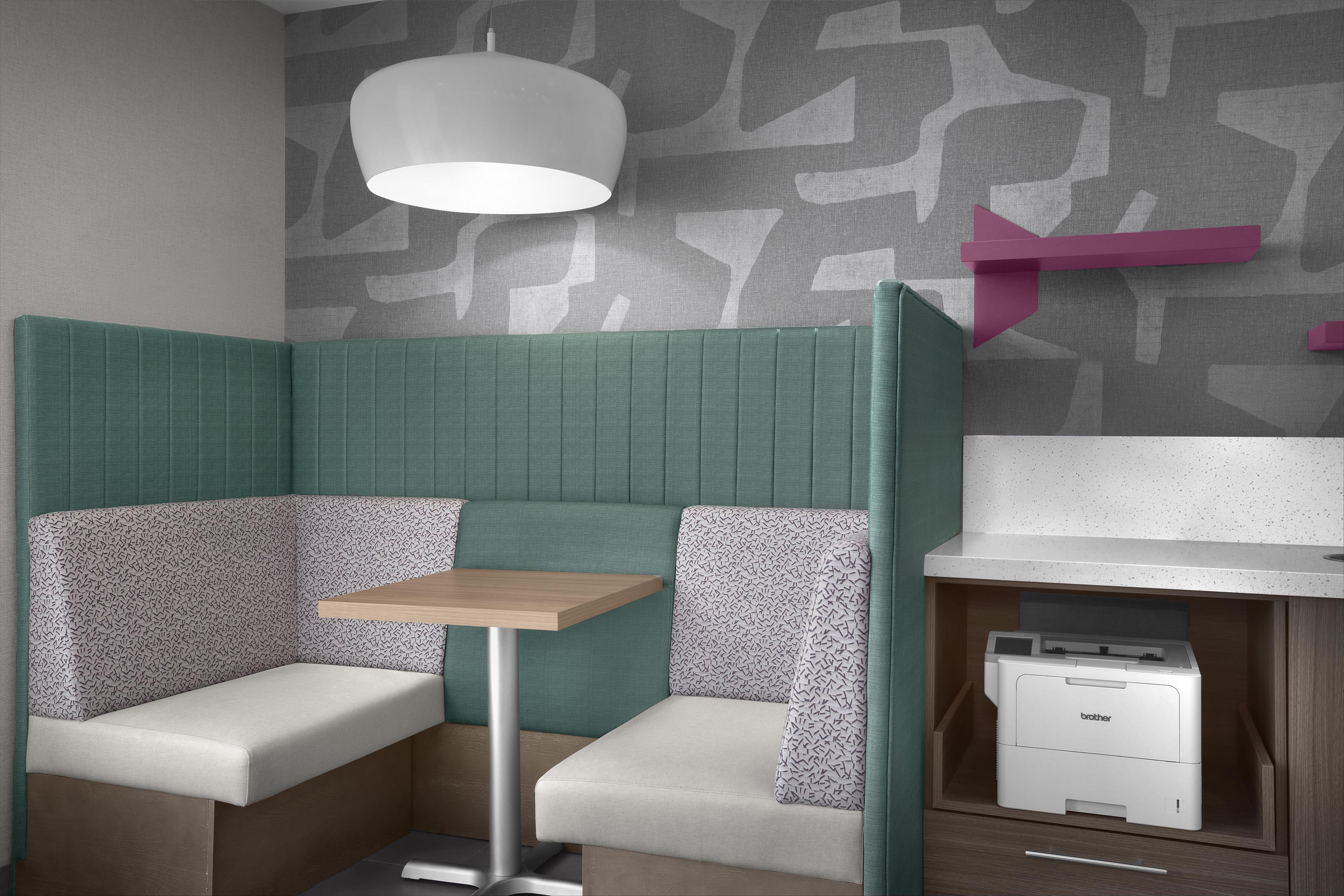 A corner booth with a table, patterned cushions, a ceiling lamp, a wall-mounted printer, and a patterned wall.