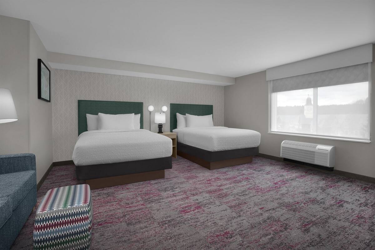 A hotel room with two beds, a window with a view, a heater, and a patterned carpet.