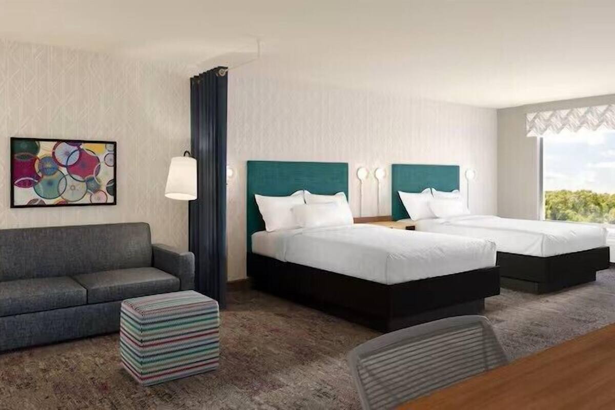A hotel room with a grey sofa, two single beds with teal headboards, a colorful abstract painting, a striped ottoman, and a view of greenery through the window.