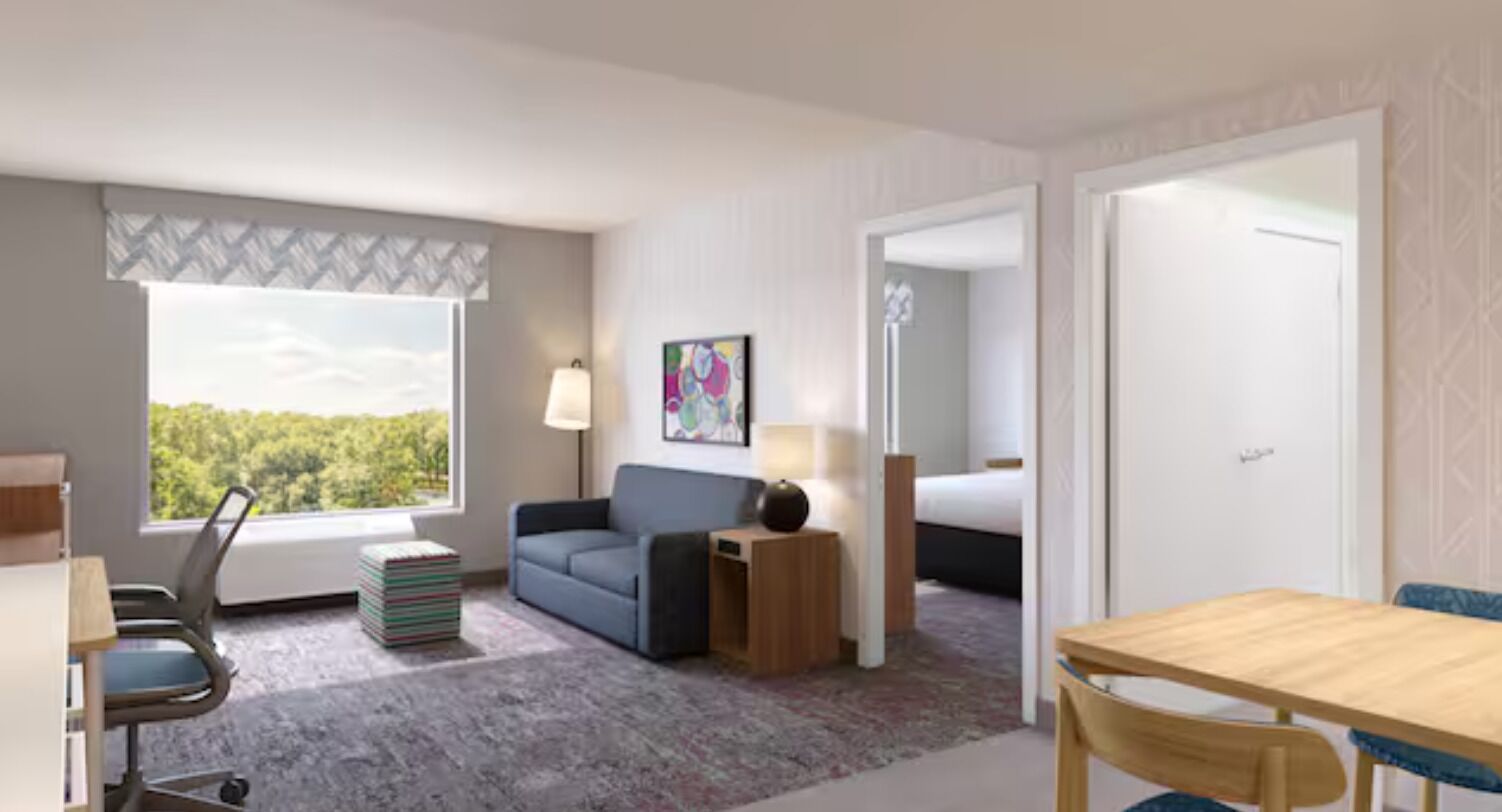 A hotel room with a desk, a chair, a sofa, a lamp, a bedside table, a bed, and a window with a view of trees.
