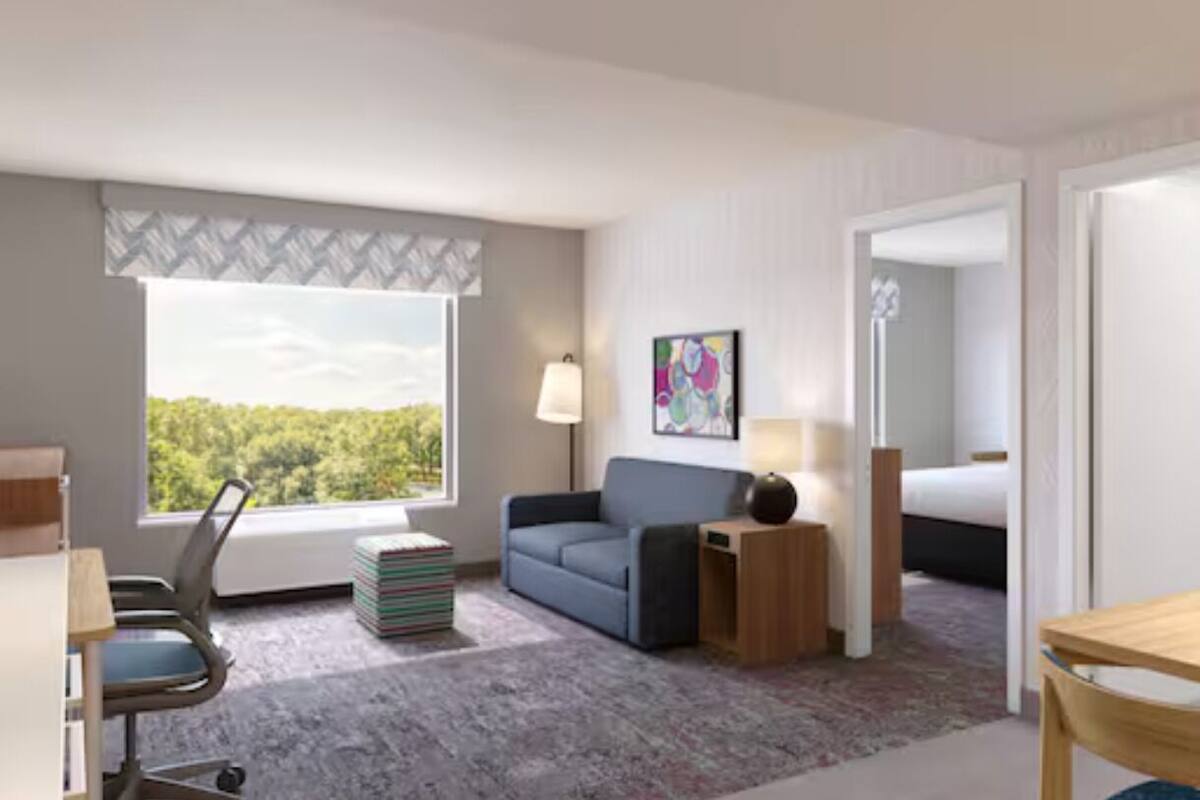 A hotel room with a desk, a chair, a sofa, a lamp, a bedside table, a bed, and a window with a view of trees.