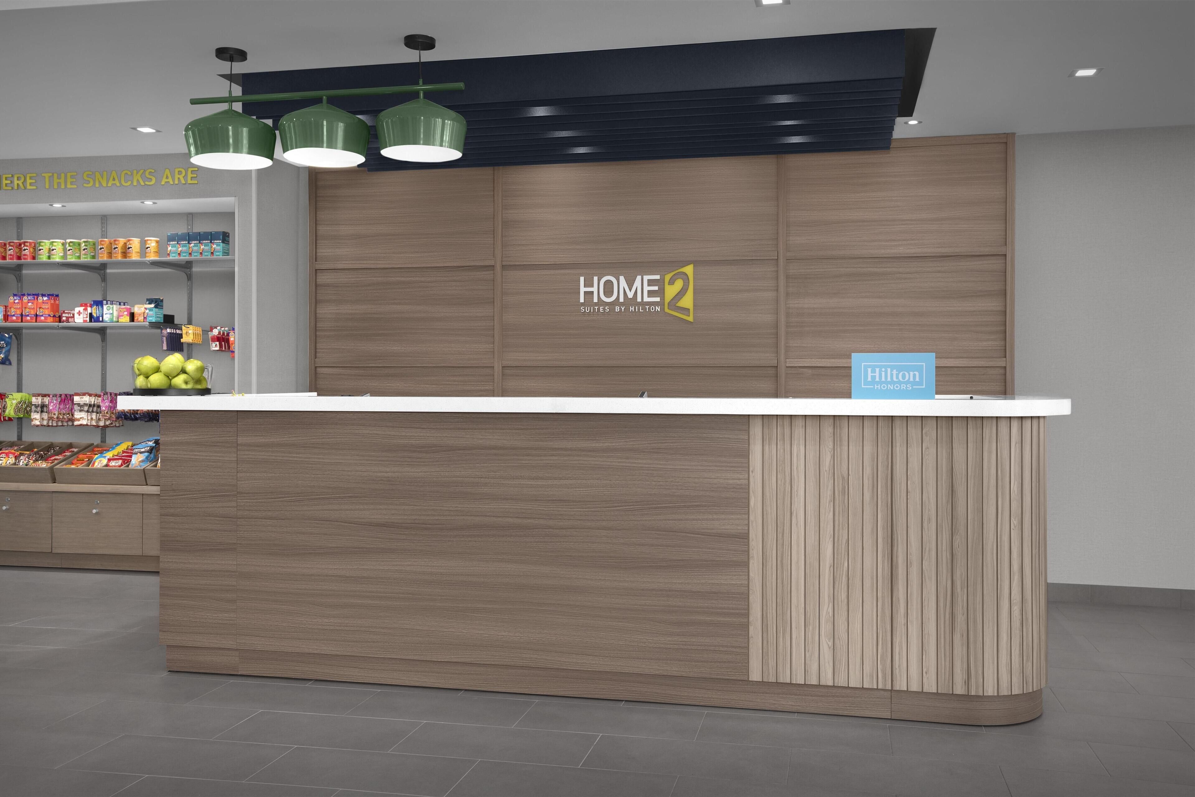 A modern hotel reception area with a wooden counter, a sign reading 'HOME 2 SUITES BY HILTON,' and a snack shelf.