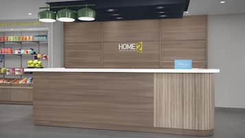A modern hotel reception area with a wooden counter, a sign reading 'HOME 2 SUITES BY HILTON,' and a snack shelf.