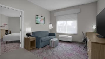 A hotel room with a bed, a blue sofa, a small ottoman, a desk with a TV, a window with blinds, and a painting on the wall.