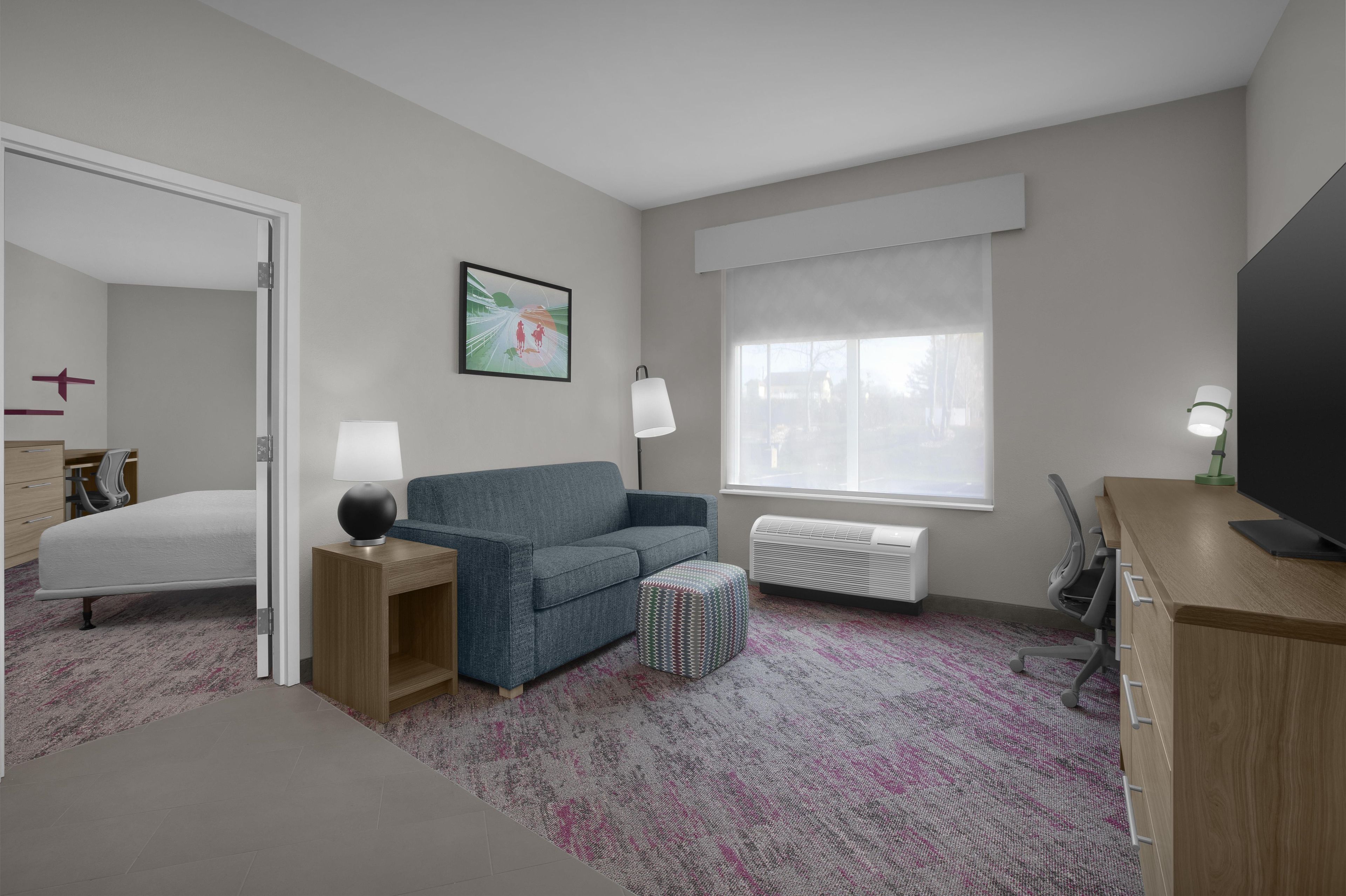 A hotel room with a bed, a blue sofa, a small ottoman, a desk with a TV, a window with blinds, and a painting on the wall.