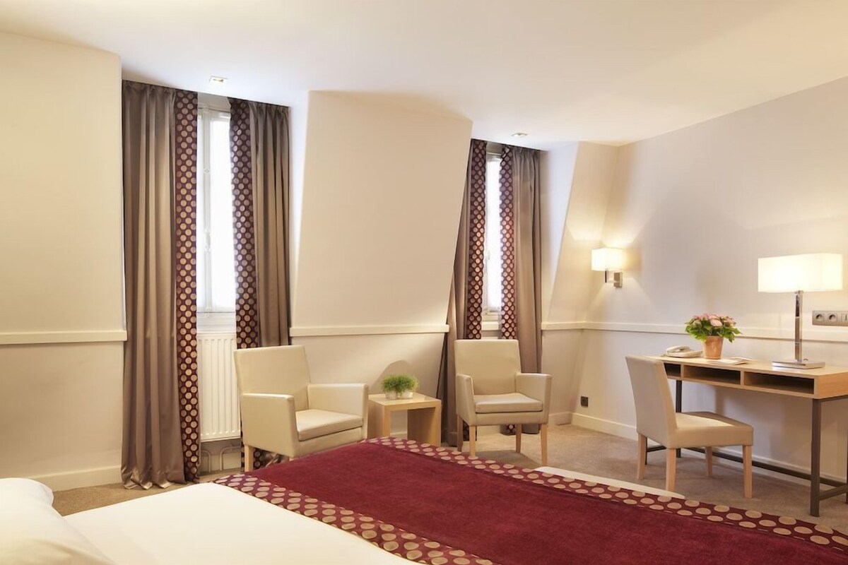 A hotel room with a bed, two chairs, a desk, a lamp, and curtains.