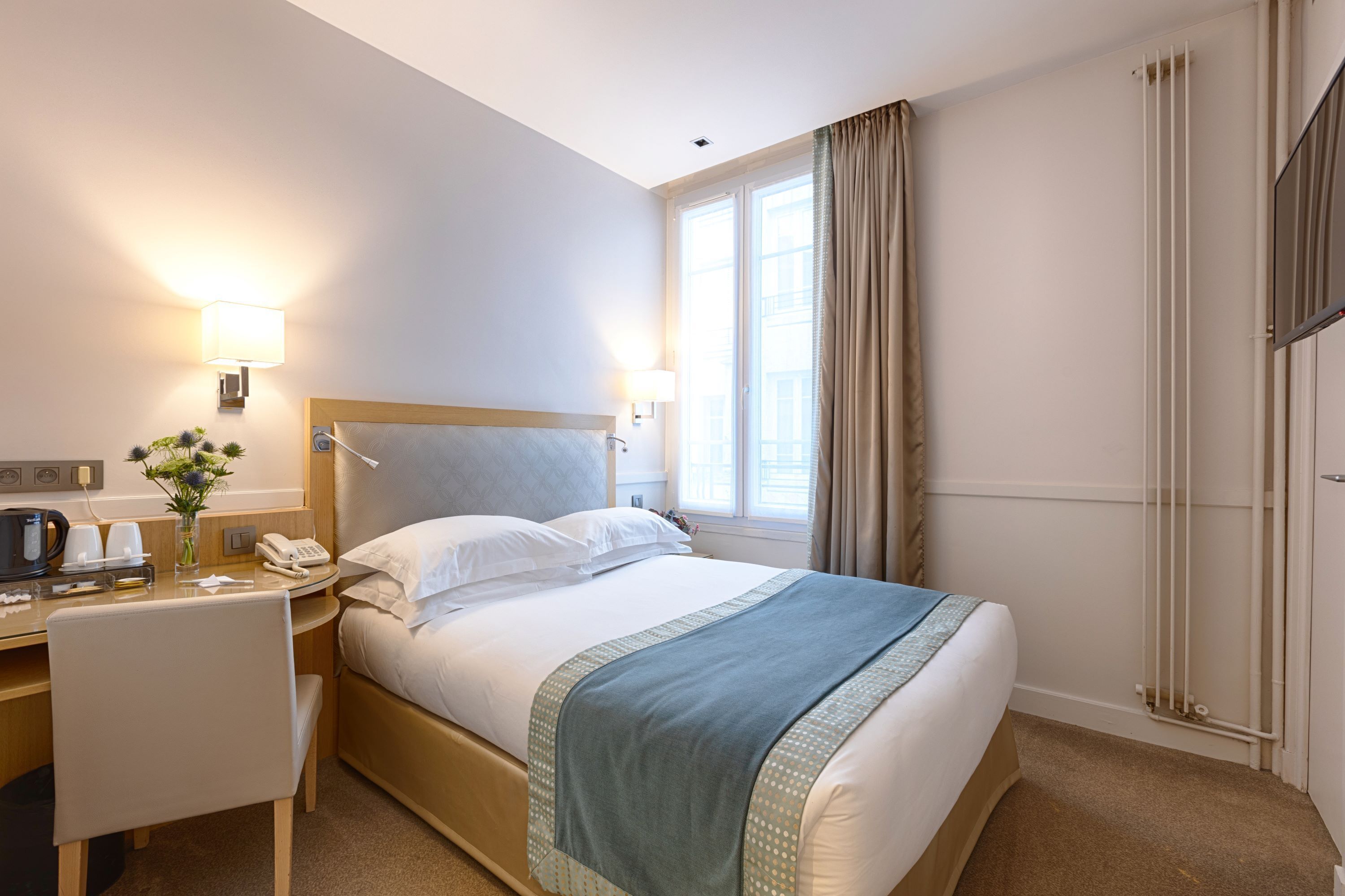 A hotel room with a bed, bedside table, lamp, window, curtains, and a desk with a telephone and coffee maker.