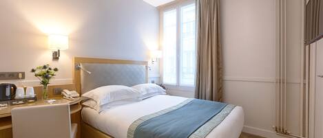 A hotel room with a bed, bedside table, lamp, window, curtains, and a desk with a telephone and coffee maker.