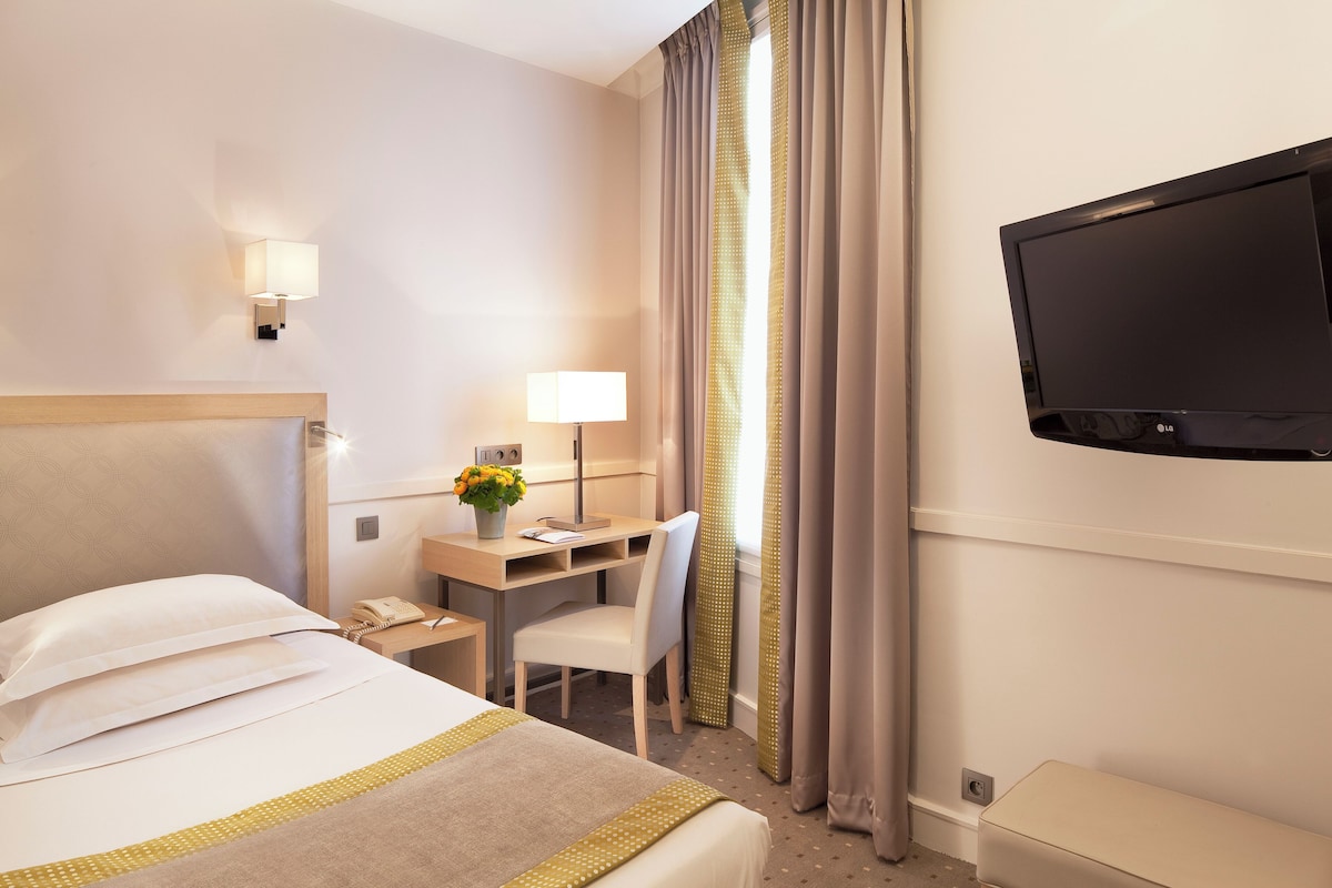 A hotel room with a bed, desk, chair, lamp, TV, and curtains.