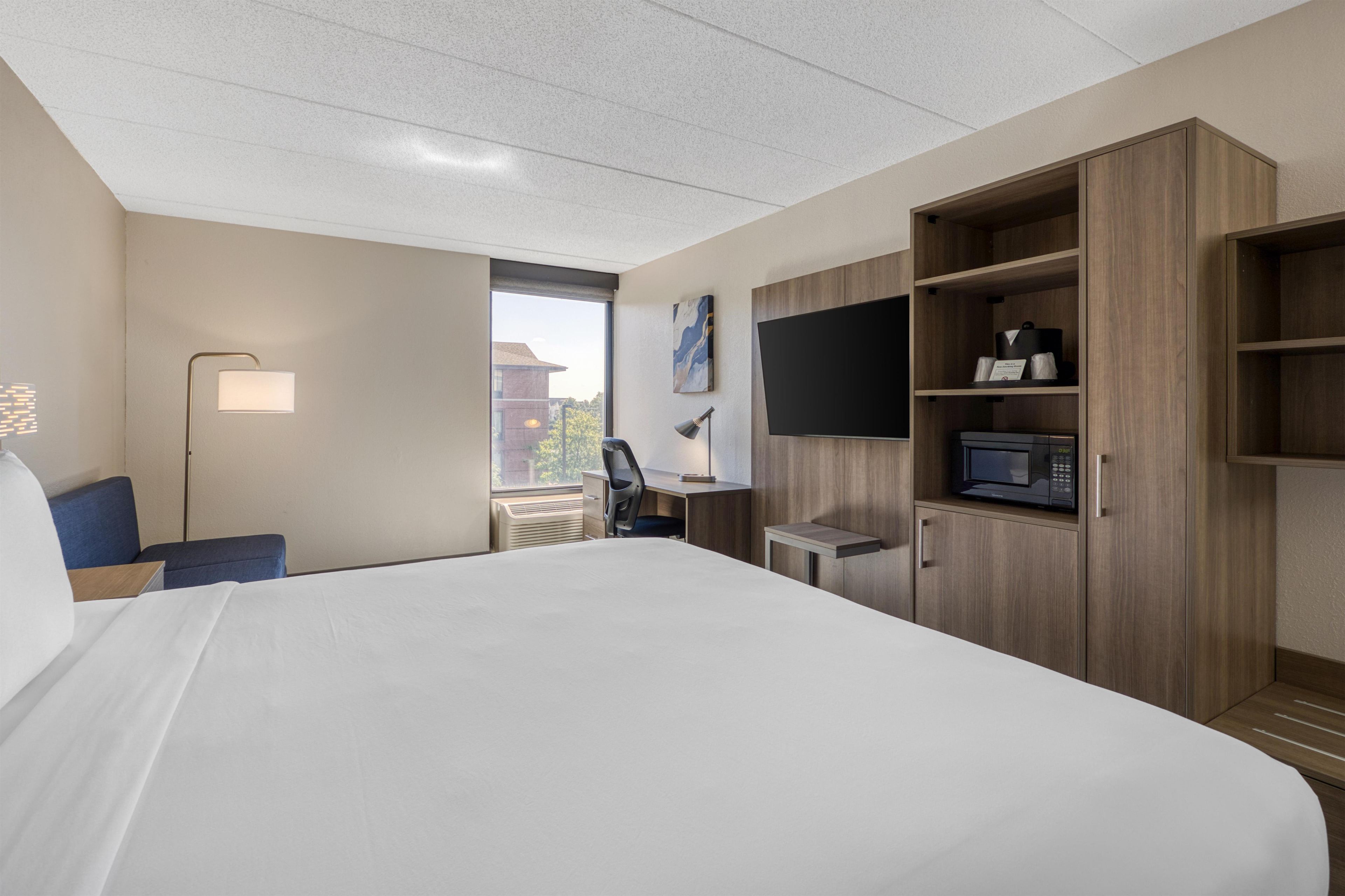 A hotel room with a bed, a desk, a chair, a TV, a microwave, and a window with a view of buildings.