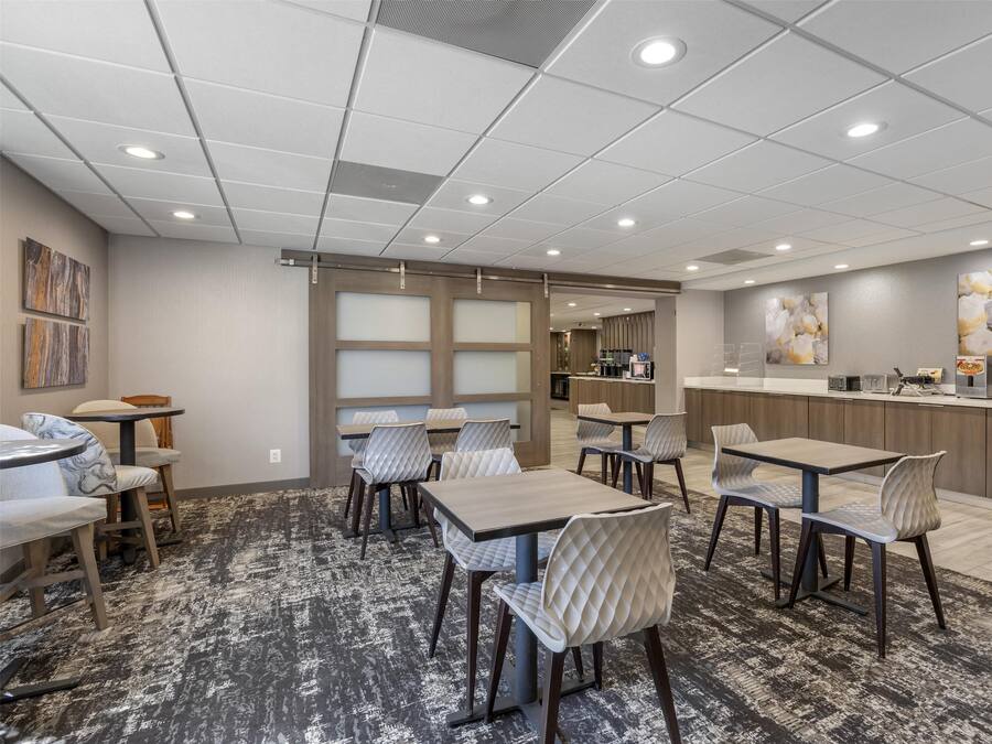 A modern hotel dining area with multiple tables and chairs, a kitchen counter, and artwork on the walls.
