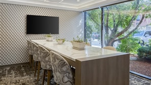 Una sala de conferencias moderna con una mesa de mármol, sillas estampadas, un televisor de pantalla plana y un amplio ventanal con vistas a árboles y coches aparcados.