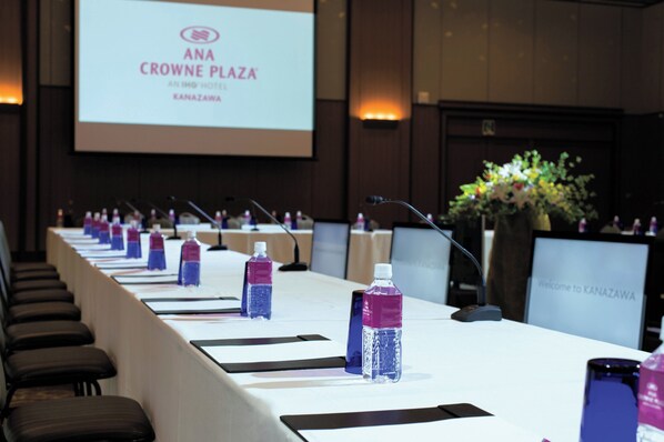 Un salón de conferencias con una larga mesa, micrófonos, botellas de agua y una pantalla que muestra 'ANA CROWNE PLAZA'.