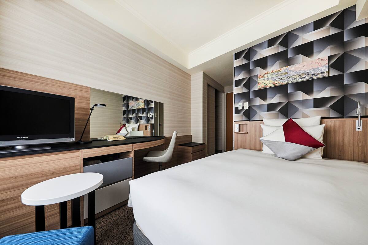 Une chambre d’hôtel moderne équipée d’un téléviseur à écran plat, d’un bureau avec une chaise, d’une table ronde, d’un lit avec des oreillers et d’un mur orné d’un motif géométrique.