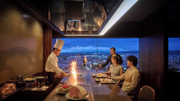 A chef cooking on an open flame with guests seated at a table with a city view.