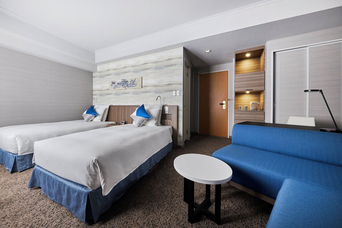 Une chambre d’hôtel avec deux lits, un canapé, une petite table et une salle de bain visible à travers une porte ouverte.