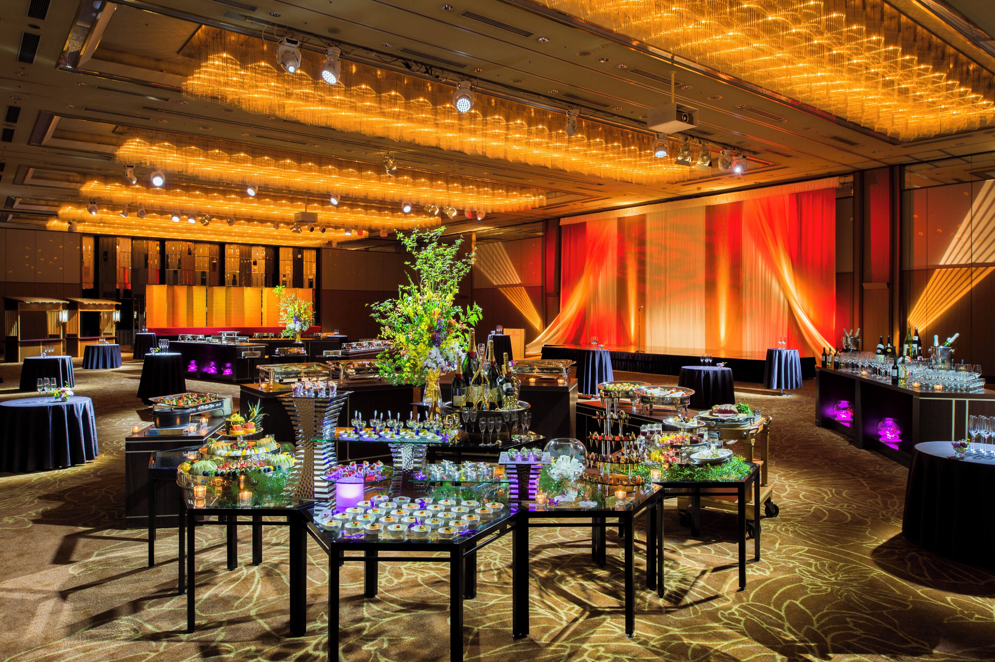 Un salón de banquetes con mesas dispuestas para un evento formal, destacando un arreglo floral central y un gran telón rojo de fondo.
