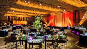 Un salón de banquetes con mesas dispuestas para un evento formal, destacando un arreglo floral central y un gran telón rojo de fondo.