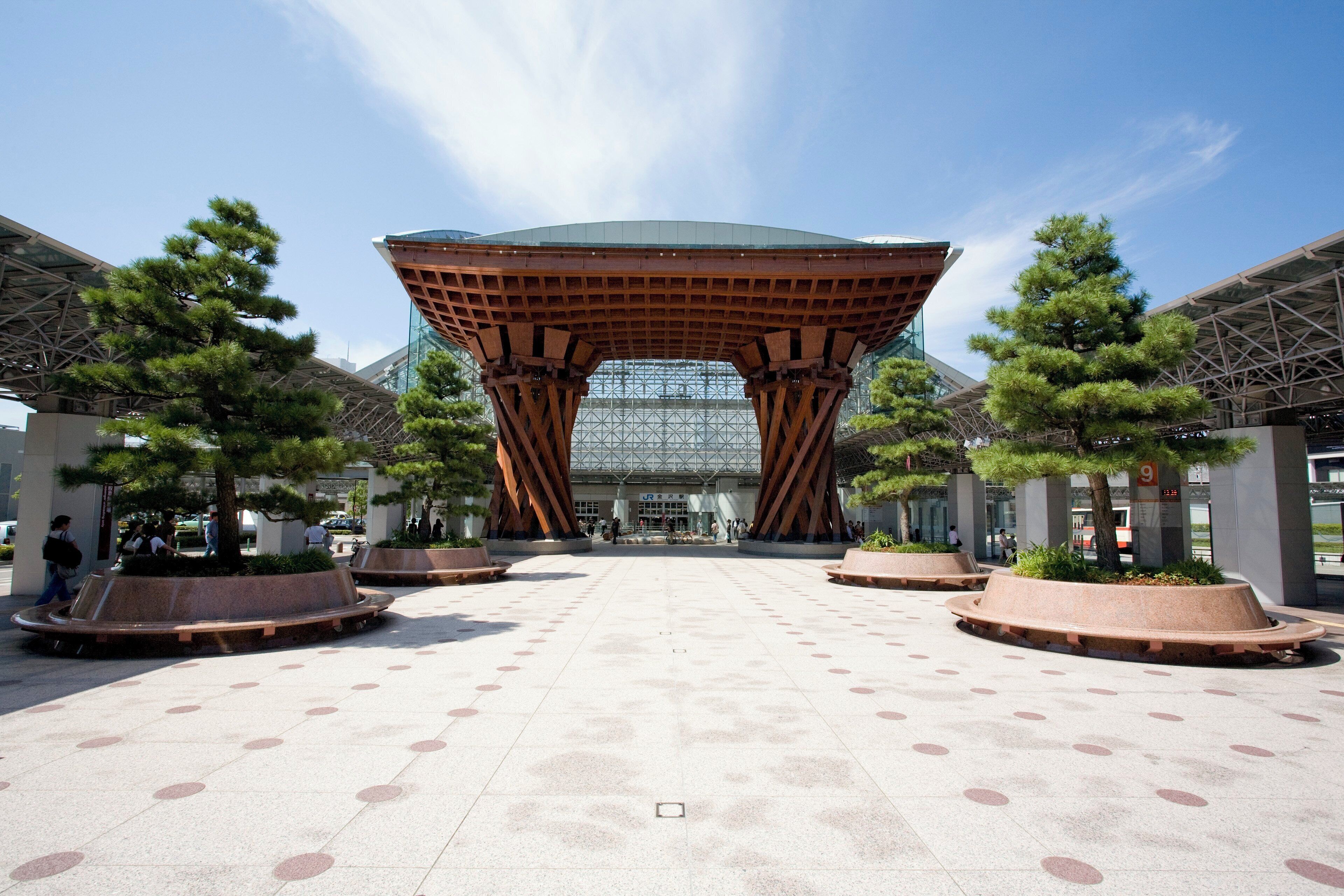 A modern building with a unique roof structure, surrounded by a plaza with patterned tiles and trees.