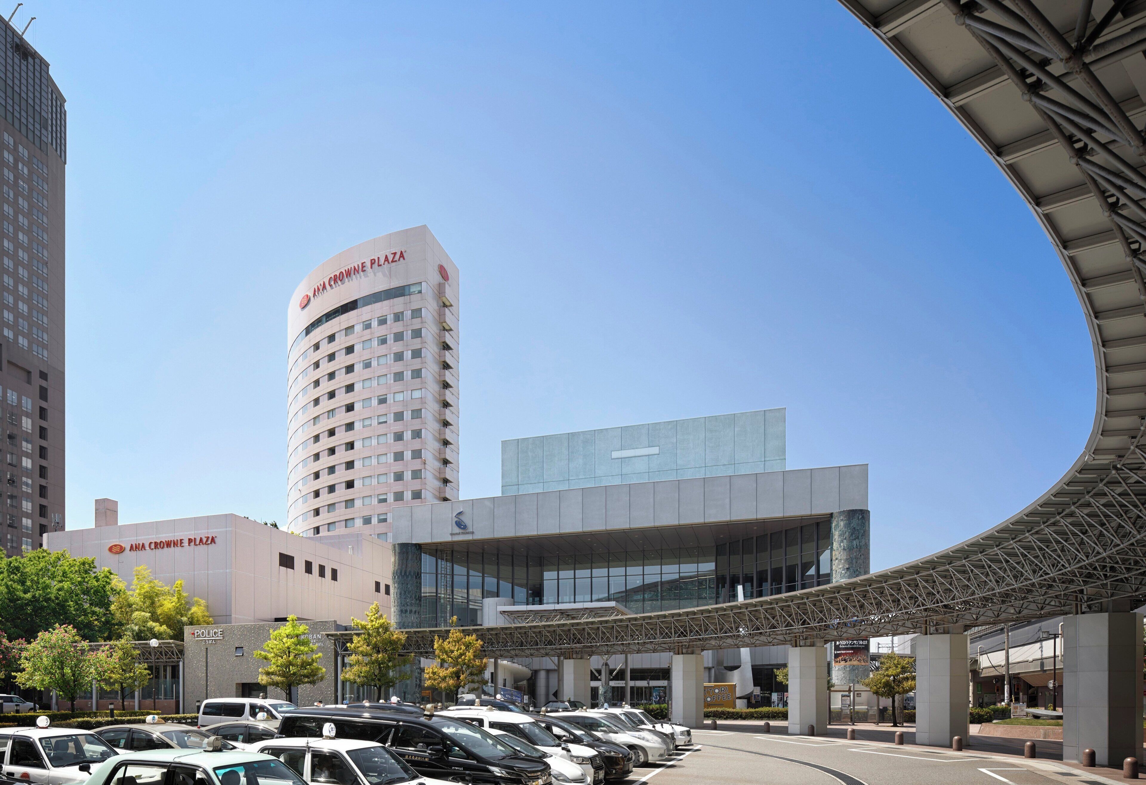 Un estacionamiento con varios autos, un edificio con la leyenda 'ANA CROWNE PLAZA' y una estructura arquitectónica moderna con techo curvo.