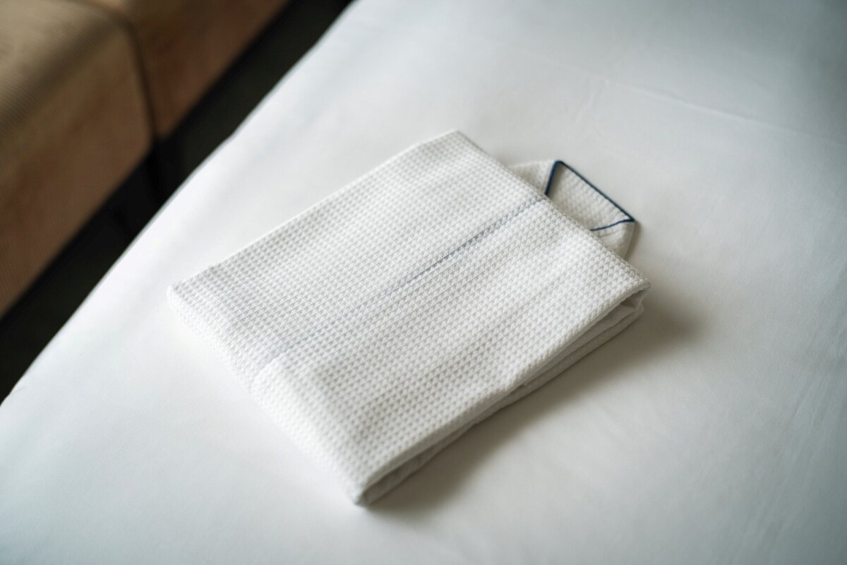 Une serviette blanche, soigneusement pliée, est posée sur un lit dont on aperçoit le cadre métallique.
