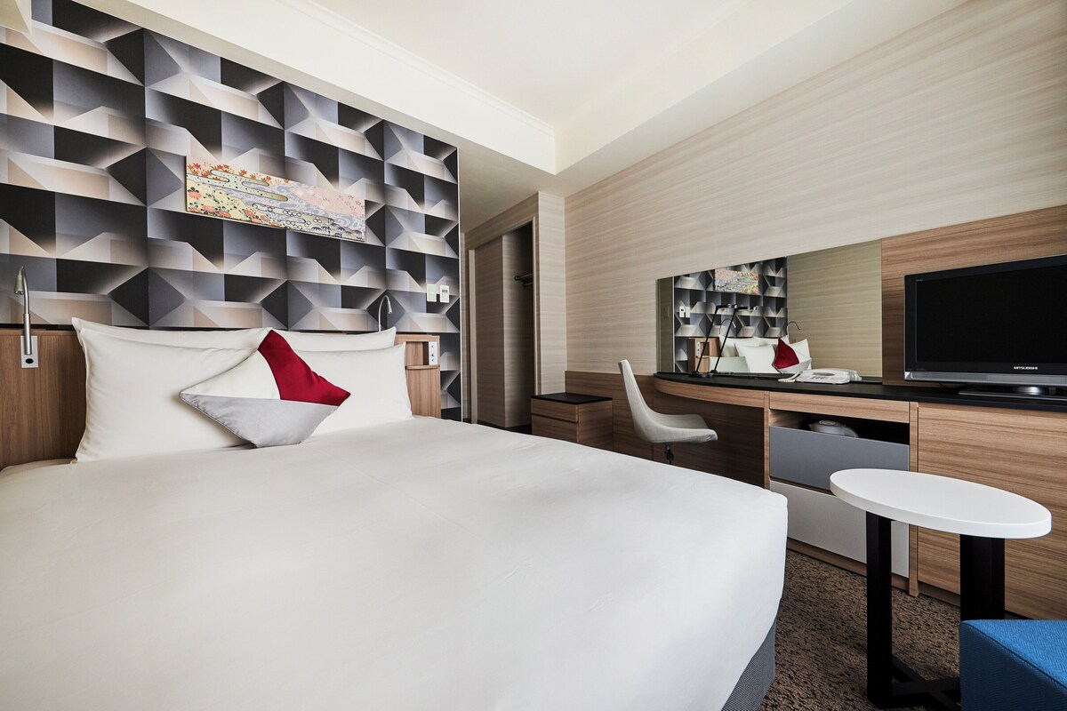 Une chambre d’hôtel équipée d’un lit, d’un bureau, d’une chaise, d’une télévision et d’un mur orné de motifs géométriques.