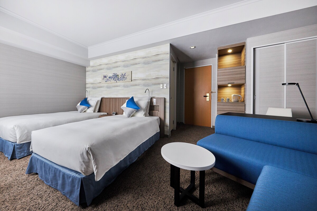 Une chambre d’hôtel avec deux lits, un canapé, une petite table et une salle de bain visible à travers une porte ouverte.